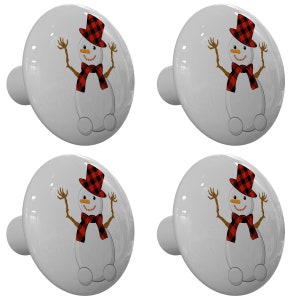 May include: Set of four white ceramic cabinet knobs with a snowman design. Each knob features a snowman with a red and black plaid hat and scarf, and a carrot nose. The snowman is waving with its arms outstretched.