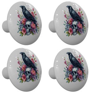 May include: Four white cabinet knobs with a decorative design. Each knob features a black raven perched above a skull, surrounded by pink, purple, and blue flowers. The knobs have a glossy finish and a rounded shape.