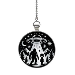 May include: A black and white pendant featuring a UFO, Bigfoot, and an alien. The design includes a flying saucer, mountains, trees, stars, and a crescent moon. The pendant hangs from a silver chain.