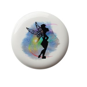 May include: A white ceramic button with a silhouette of a fairy with iridescent wings against a watercolor background of blue, pink, and purple.