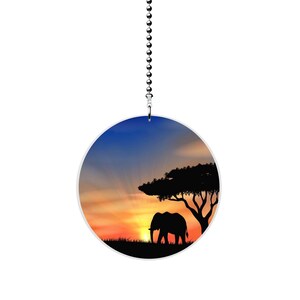 May include: A round ceiling fan pull with a silhouette of an elephant walking under a tree at sunset. The sky is a gradient of blue, orange, and yellow.