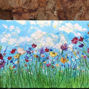 May include: An oil painting of a vibrant wildflower meadow under a bright blue sky with fluffy white clouds. The artwork features a variety of colorful flowers in shades of red, purple, yellow, and white, set against a backdrop of green grass.