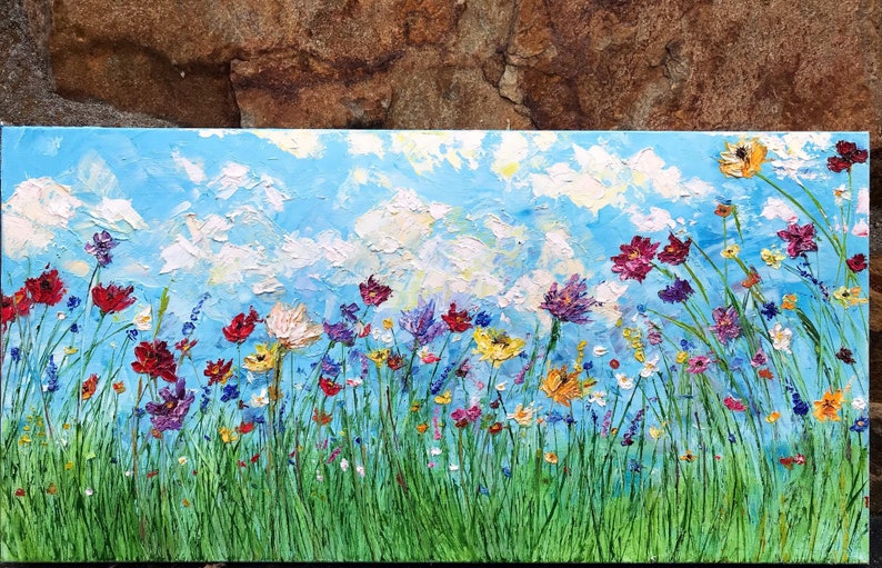 May include: An oil painting of a vibrant wildflower meadow under a bright blue sky with fluffy white clouds. The artwork features a variety of colorful flowers in shades of red, purple, yellow, and white, set against a backdrop of green grass.