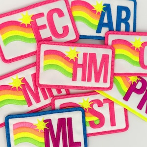 Könnte beinhalten: Eine Sammlung bunter gestickter Patches mit Regenbogen-Designs und verschiedenen Buchstaben. Die Patches sind rosa, blau, gelb und grün mit weißen Hintergründen. Die Buchstaben auf den Patches sind EC, AR, HIM, ML, ST und PR.