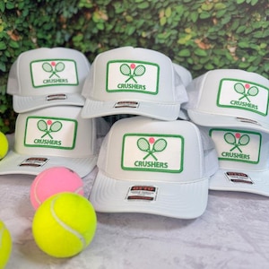 Op de afbeelding: Een verzameling witte truckerpetten met een rechthoekige patch met een groene rand, twee gekruiste tennisrackets, een kleine roze cirkel en de tekst CRUSHERS. Twee tennisballen, één neongeel en één roze, liggen op de voorgrond.