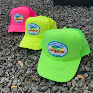 May include: Three trucker hats in pink, yellow and neon green with a palm tree logo and the text "PortATX 2022 Est. 2014".