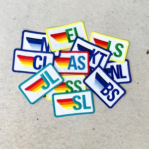 May include: A collection of colorful iron-on patches with rainbow stripes and letters spelling out "AS", "BS", "CL", "EL", "JL", "KT", "NL", "SL", "SS" and "IS".