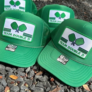 May include: Three green trucker hats with a white patch featuring a green and blue paddleball design and the text "TEAM DOUBLE D".