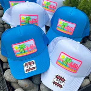 May include: Several trucker hats in shades of blue and white, with a mesh back. Each hat has a rectangular patch with a tropical design, including palm trees, a sun, and initials. The hats have the text "OTTO WORLD FAMOUS FOAM FRONT OSFM TRUCKER".