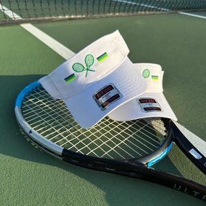 May include: Two white tennis visors with embroidered green tennis racket designs and colourful stripes. The visors rest on a blue and black tennis racket on a green court, with a net in the background.