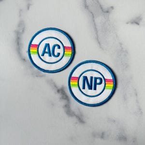 Op de afbeelding: Twee ronde geborduurde patches met een regenboogstreepontwerp. De eerste patch is blauw met witte letters die "AC" zeggen. De tweede patch is blauw met witte letters die "NP" zeggen.