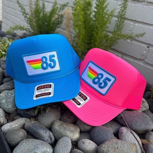 May include: Two trucker hats with a rainbow stripe and the number 85 on the front. One hat is blue and the other is pink.