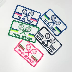 Puede incluir: Parches rectangulares bordados con gráficos de raquetas de tenis y texto. Los parches presentan frases como "I'd Hit That" y "Over-Served" en varios colores, incluyendo rosa, verde y azul. Los parches son blancos con bordes de colores.