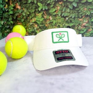 Op de afbeelding: Een witte zonneklep met een geborduurde patch van twee gekruiste tennisrackets en een kleine roze bal. De klep heeft een zwart-zilveren sticker op de rand met de tekst OTTO CLASSIC FIT SUN VISOR. Verschillende gele tennisballen.