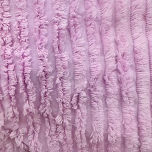May include: Close-up of a soft, plush fabric with a light purple color and a textured, fuzzy surface. The fabric has a vertical striped pattern created by rows of small, fluffy tufts.