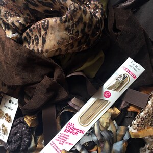 May include: A close-up of a variety of fabric scraps, including leopard print, brown velvet, and sequins. A package of a 7 inch, 18 cm, camel colored zipper is visible in the center of the image. The package is labeled "All Purpose Zipper" and "Polyester".
