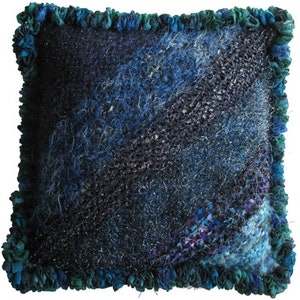 Puede incluir: Cojín cuadrado decorativo con un diseño tejido a mano. Presenta un degradado de azules y tonos oscuros, con un borde verde con volantes. El patrón tejido crea una estética única y acogedora, adecuada para la decoración del hogar.