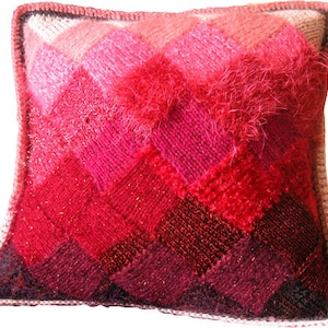 Puede incluir: Un cojín decorativo cuadrado con un diseño de patchwork en tonos rojos, rosas y burdeos. El cojín presenta un patrón de punto texturizado con una sección esponjosa y peluda en el centro. Los bordes están acabados con un borde de ganchillo.