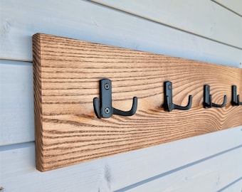 Könnte beinhalten: Ein Wand-Kleiderständer aus Holz mit sechs schwarzen Metallhaken. Der Kleiderständer ist dunkelbraun gebeizt und hat eine natürliche Holzmaserung.