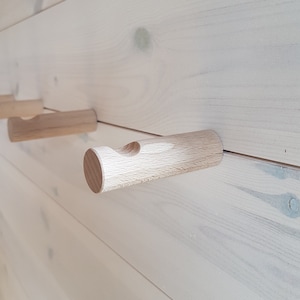 Può includere: Ganci appendiabiti in legno montati su una parete di legno di colore chiaro. I ganci sono cilindrici con un'incavatura curva per appendere oggetti. Il legno è di colore marrone chiaro naturale e la parete ha una finitura sottile di colore chiaro.