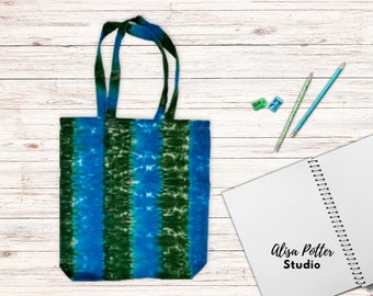 May include: A blue and green tie-dye tote bag with a white background. The bag has a wide, flat bottom and two long handles. The bag is sitting on a white wooden surface.