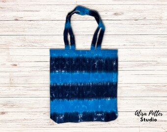 May include: A blue and black tie-dye tote bag with a white background. The bag has two straps and is made of canvas.