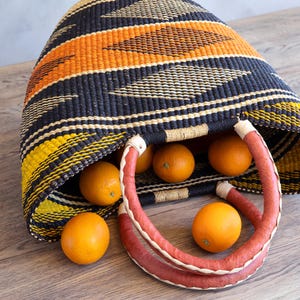 Könnte beinhalten: Ein geflochtener Korb mit einem geometrischen Muster in Schwarz, Orange und Gelb. Der Korb hat zwei abgerundete Griffe, die mit einem strukturierten, rotbraunen Material umwickelt sind. Mehrere leuchtend orange Zitrusfrüchte liegen darin.