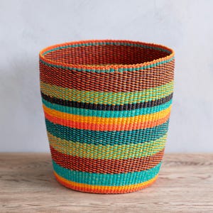 Peut inclure: Un panier cylindrique tissé avec un motif rayé vibrant. Le panier présente des bandes orange, sarcelle, jaune et noir. Le panier est fait de fibres naturelles et repose sur une surface en bois.