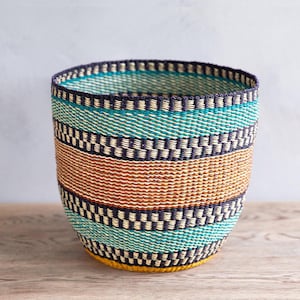 Könnte beinhalten: Ein gewebter Korb mit zylindrischer Form. Er hat horizontale Farbstreifen, darunter Türkis, Braun und Schwarz, mit einem gelben Boden. Das Design beinhaltet ein sich wiederholendes Muster, das ein strukturiertes Aussehen erzeugt. Geeignet zur Aufbewahrung oder Dekoration.