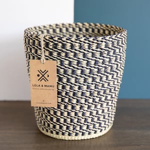 Peut inclure: Une corbeille à papier tissée en noir et naturel avec une étiquette brune sur laquelle est inscrit "Lola & Mawu Ethical African Decor lolaandmawu.com".