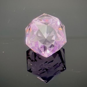 May include: A faceted, light purple gemstone with a complex geometric shape. The gem is resting on a reflective surface, creating a dark purple mirrored image below. The gem's facets catch the light, creating a sparkling effect.