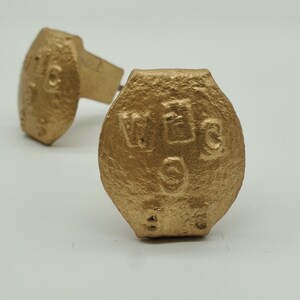 May include: Two gold-colored cabinet knobs with a textured surface. Each knob has a rectangular stem and a rounded, flattened face with embossed letters and numbers. The knobs are set against a white background.