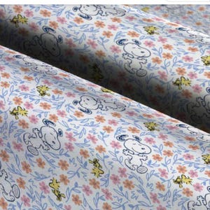 Puede incluir: Tela blanca con un patr&oacute;n repetido de Snoopy y Woodstock. Snoopy est&aacute; representado en un contorno azul y Woodstock es amarillo. El fondo es una tela blanca con un patr&oacute;n floral azul, rosa y naranja.