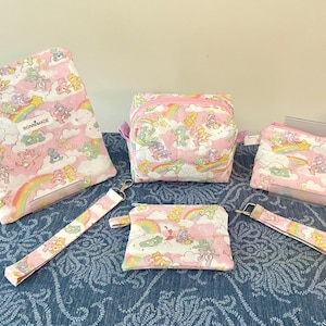May include: A collection of pink quilted accessories featuring a repeating pattern of cartoon bears, rainbows, and clouds. The set includes a large pouch, a square bag, a small zippered pouch, and two wrist straps. The text "BONNMADE" is visible on the large pouch.