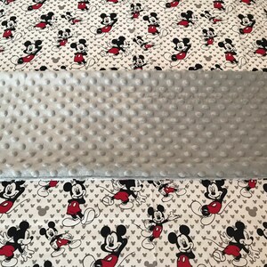 May include: A gray, textured blanket with a white pillowcase featuring a repeating pattern of Mickey Mouse in red, black, and white. The pillowcase also has gray heart shapes.