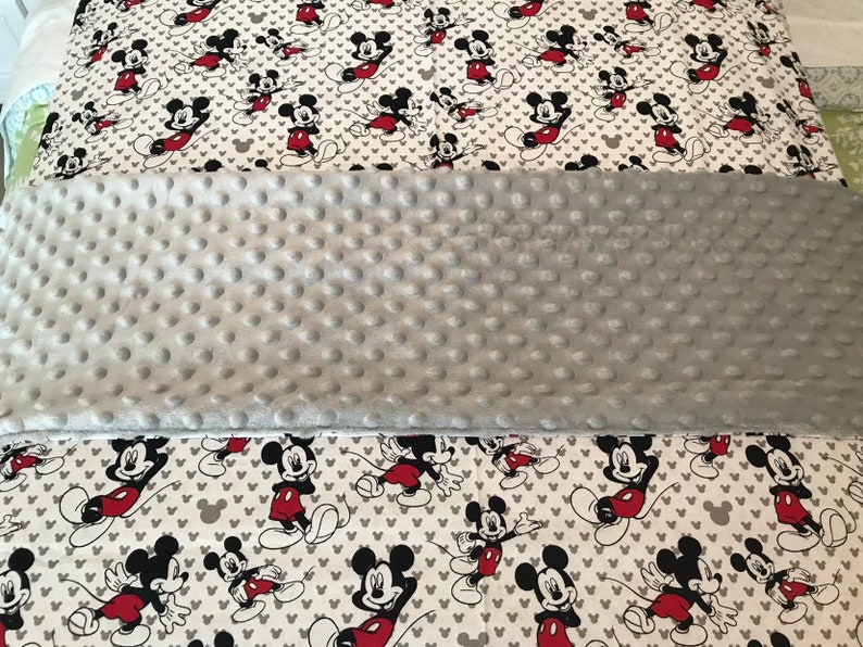 May include: A gray, textured blanket with a white pillowcase featuring a repeating pattern of Mickey Mouse in red, black, and white. The pillowcase also has gray heart shapes.