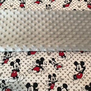 May include: A baby blanket featuring a gray, dimpled, plush fabric and a white fabric printed with Mickey Mouse in various poses. Mickey is wearing red shorts and black shoes. The background has gray Mickey Mouse head silhouettes.