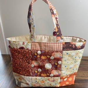 Op de afbeelding: Een patchwork draagtas met diverse herfstachtige stofpatronen, waaronder pompoenen, wilde bloemen en oranje gingham. De tas heeft twee stoffen handvatten en een crèmekleurige voering aan de binnenzijde, afgewerkt met zichtbare stiksels.