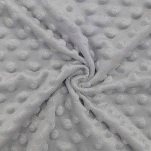 May include: Close-up of a soft, light gray fabric with a textured, dimpled surface. The fabric is folded and swirled, showing the plush texture and the raised, rounded dots that cover the material. The fabric appears to be a plush, cozy material.