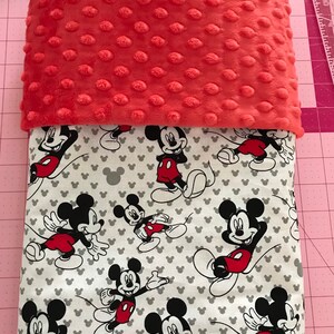 May include: A coral-colored, textured blanket top over a white blanket with a Mickey Mouse pattern. The Mickey Mouse pattern features the iconic character in various poses, with red shorts and black gloves.