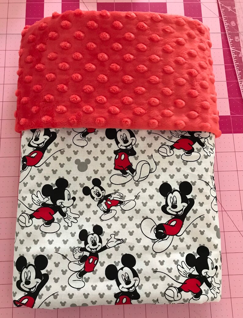 May include: A coral-colored, textured blanket top over a white blanket with a Mickey Mouse pattern. The Mickey Mouse pattern features the iconic character in various poses, with red shorts and black gloves.