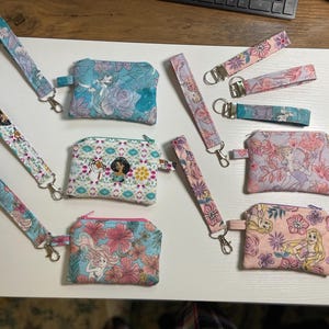 Könnte beinhalten: Eine Sammlung von Stoff-Handgelenkschlaufen und kleinen Reißverschlusstaschen. Die Handgelenkschlaufen und Taschen weisen Blumen- und Charakterdesigns in Blau-, Rosa- und Weißtönen auf. Einige haben einen Metallverschluss.
