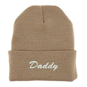 May include: A tan beanie hat with a folded brim. The word "Daddy" is embroidered in a cursive font in white thread on the front of the hat. The hat is made of a ribbed knit material.