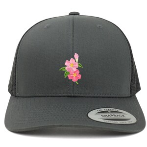 Puede incluir: Una gorra de camionero gris oscuro con un bordado de flores rosas en la parte delantera. La gorra tiene una parte trasera de malla negra y un cierre de presión negro. La gorra tiene una pegatina en la parte trasera que dice "The Classics Snapback Yupoong Since 1974".