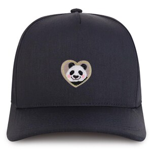 Könnte beinhalten: Marineblaue Baseballkappe mit einem gestickten Panda-Bär-Design in einem herzförmigen Aufnäher. Der Panda hat schwarz-weißes Fell mit einem rosa Rouge. Das Herz ist goldfarben umrandet.