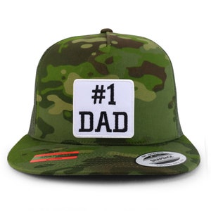 Op de afbeelding: Een groene camouflage trucker cap met een wit vierkant patch waarop in zwarte letters "#1 DAD" staat.