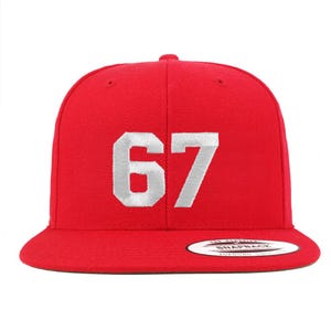 Puede incluir: Gorra snapback roja con el número "67" bordado en blanco en la parte delantera. La gorra tiene una visera plana y una corona estructurada. La gorra es de color rojo liso.
