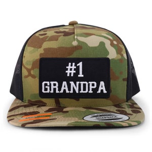 Op de afbeelding: Snapback-pet met camouflagepatroon en een zwarte mesh-achterkant. De pet heeft een zwart rechthoekig embleem met de tekst "#1 GRANDPA" in het wit geborduurd. De pet heeft een gebogen klep en een bruine knoop aan de bovenkant.
