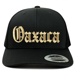 May include: Black snapback hat with the word "Oaxaca" embroidered in gold gothic lettering. The hat features a mesh back and a flat brim. The tag on the brim reads "The Classics Snapback".