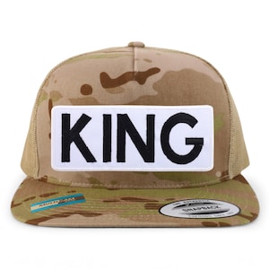 Puede incluir: Una gorra de camionero de camuflaje marrón y verde con un parche blanco que dice "KING" en letras negras.